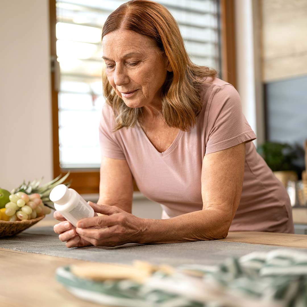 An older woman reads the label on an over-the-counter medication bottle in her kitchen.]